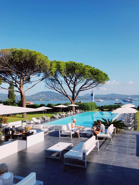 A poolside lounge area with white furniture, overlooking a scenic bay and hills on a sunny day.